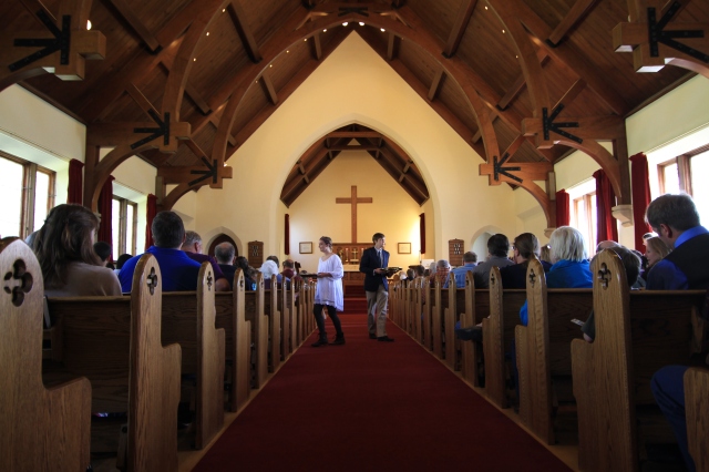 Photo of the interior of the Mammoth Chapel. Two ushers on a red carpet hold tithe plates. Parishioners sit in pews.